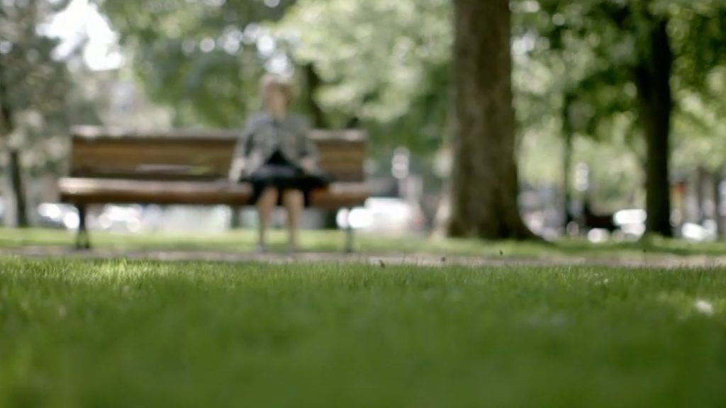 Image floue : Nicole assise sur un banc dans un très beau parc, verdoyant et lumineux