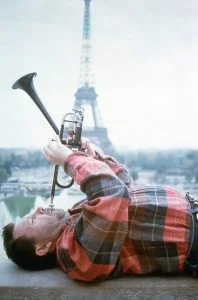 Photo de Maurice Gourgues, couché sur le dos, jouant de la trompette, avec la Tour Eiffel en arrière plan.